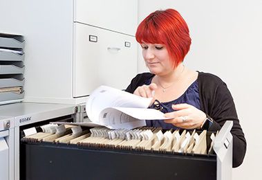 Mirjam Nagl standing behind a filing cabinet. She is reading a document. (photo)