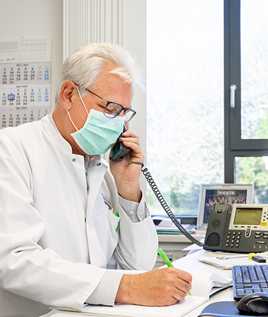 Site physician Dr. Wolfgang Großwieser having a phone call at a desk. He is wearing a face mask. (Photo)